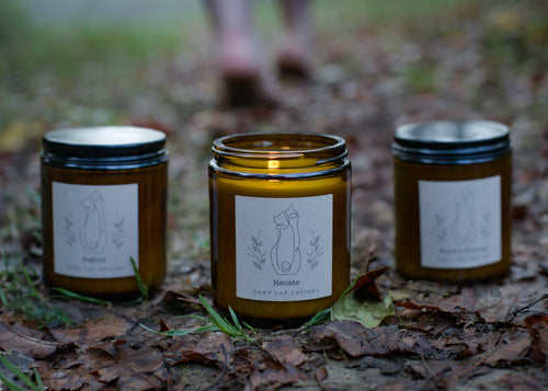 Three candles in amber jars with labels on a leaf-covered ground